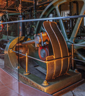 Historic industrial museum with a massive steam engine and flywheel behind glass, illuminated by warm orange lighting.