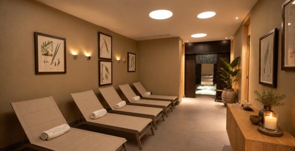 Warmly lit spa relaxation room with chaise lounges, rolled towels, botanical art, plants, and candles.