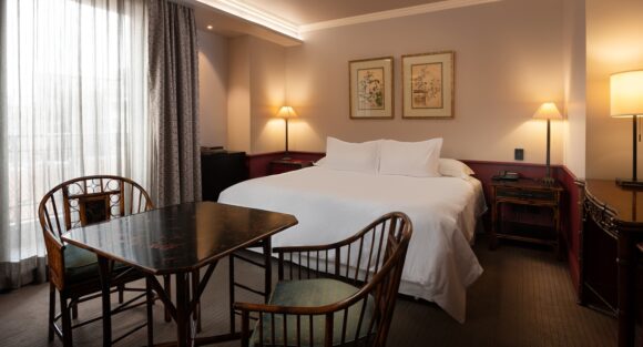 Elegant hotel room with a king bed, antique dining table and chairs, framed art, warm lamps, and sheer-curtained windows.