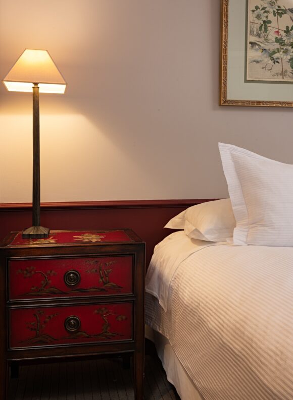 Cozy bedroom with a glowing table lamp on an ornate red bedside chest beside a neatly made bed and framed floral art.