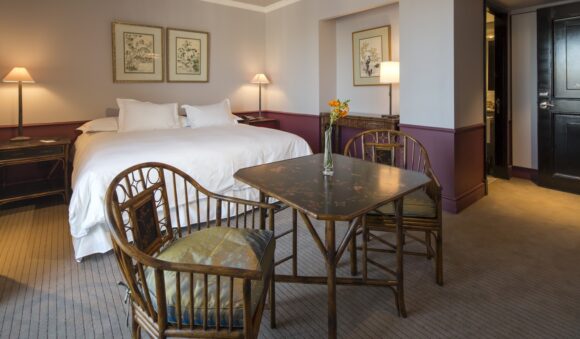 Elegant hotel bedroom with a white bed, antique wooden dining table and chairs, framed botanical art, and warm lamplight.