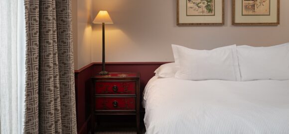 Elegant bedroom with a white bed, red bedside table, warm floor lamp, patterned curtains, and framed botanical art.