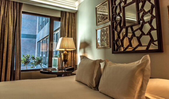 THE LASTARRIA ROOM_Lastarria Room (1)-RET Elegant hotel bedroom with white pillows, a brass bedside lamp, city-view window, and geometric wood-framed mirror.
