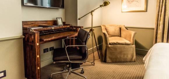 THE LASTARRIA ROOM_Lastarria Room (3) Hotel room desk with wooden writing desk, office chair, wall-mounted TV, floor lamp, upholstered armchair, and framed artwork.