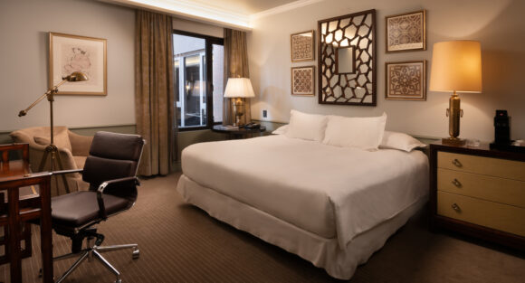 THE LASTARRIA ROOM_NZ6_8738-2-Edit_V4 Elegant hotel bedroom with a king bed, warm lighting, framed geometric art, a desk, and a seating area.