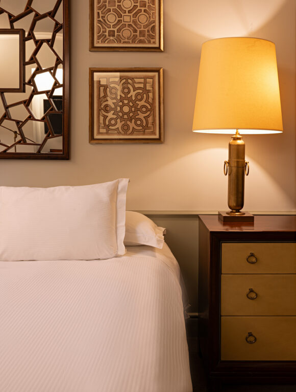 THE LASTARRIA ROOM_NZ6_8807-2-Edit_V3_sh Cozy bedroom with white bed, glowing brass table lamp, geometric framed artwork, and dark wood nightstand beside a decorative mirror.