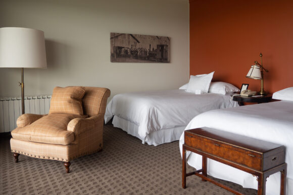 Hotel room with two white beds, a tan armchair, bedside lamps, antique wooden luggage bench, and rust-red accent wall.