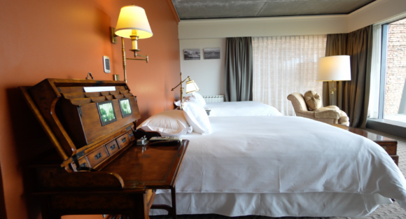 Elegant hotel bedroom with two white beds, antique wooden desk, warm lamps, and a lounge chair beside large curtained windows.