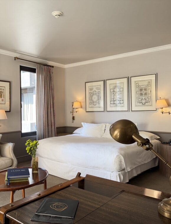 Elegant hotel suite with a king bed, cream sofa, framed architectural prints, tall curtained windows, and brass lighting.