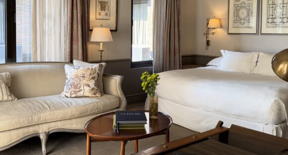 Elegant hotel bedroom with white bed, cream sofa, framed architectural prints, tall curtained windows, and a flower vase on a coffee table.