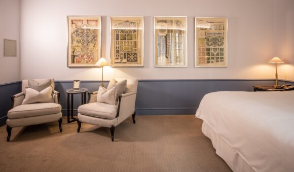 Elegant hotel bedroom with a white bed, two upholstered armchairs, warm lamps, and framed vintage garden plans on the wall.