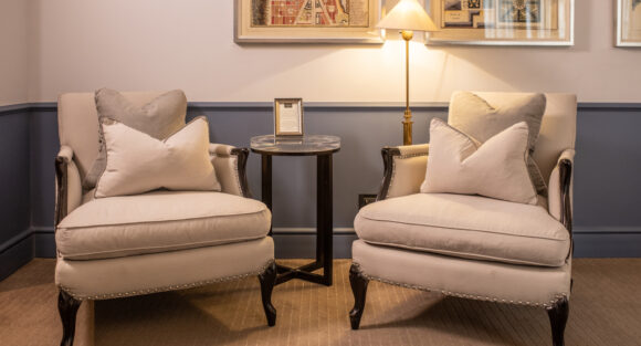 Two cream armchairs with pillows flank a round table beneath framed vintage maps and a glowing floor lamp.