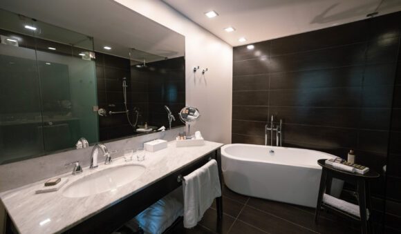 THE SINGULAR KING_baño2 Modern hotel bathroom with marble vanity, wall-sized mirror, glass shower, freestanding bathtub, and dark tile walls.