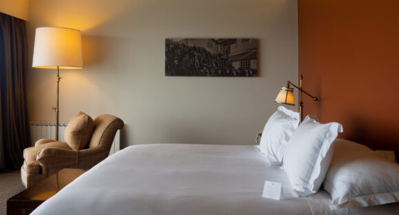 THE SINGULAR KING_king3 Warmly lit hotel bedroom with a white bed, brass reading lamp, plaid armchair, and historic black-and-white wall photograph.