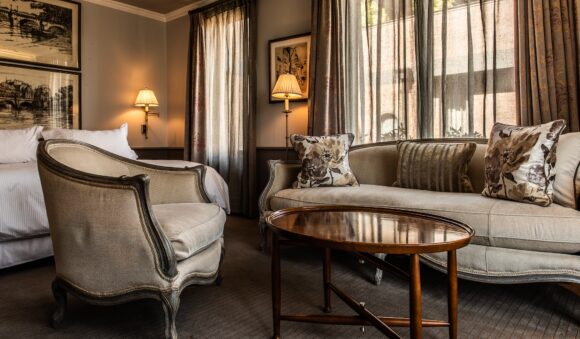 Elegant hotel suite with a bed, upholstered sofa and armchair, oval coffee table, framed art, and sunlit draped windows.