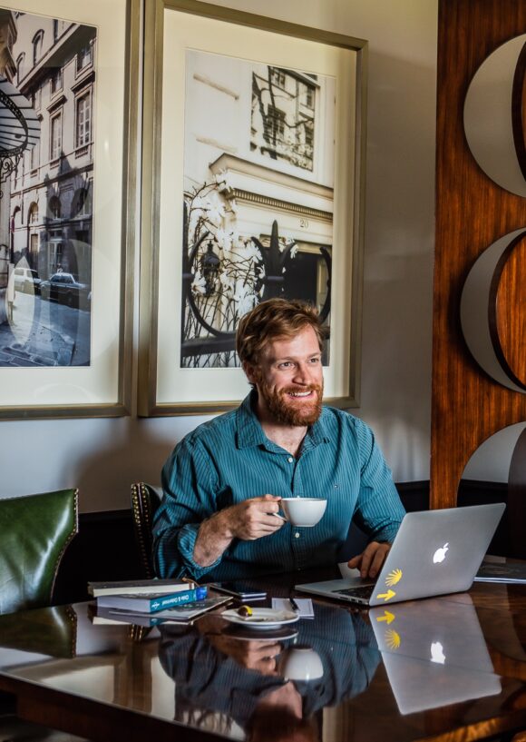 Smiling bearded man works on a laptop while holding a coffee cup at a table in a stylish lounge.