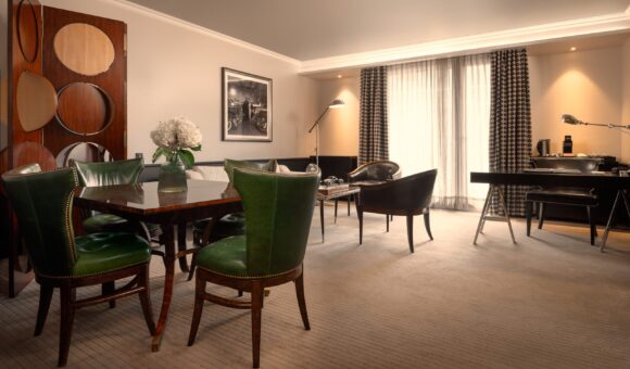 Elegant hotel suite with green leather dining chairs, a polished wood table, lounge seating, and a desk beside large curtained windows.