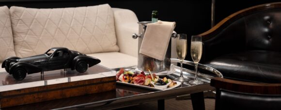 Elegant lounge with cream sofa, black leather chair, champagne flutes, silver ice bucket, appetizer platter, and model car on a table.
