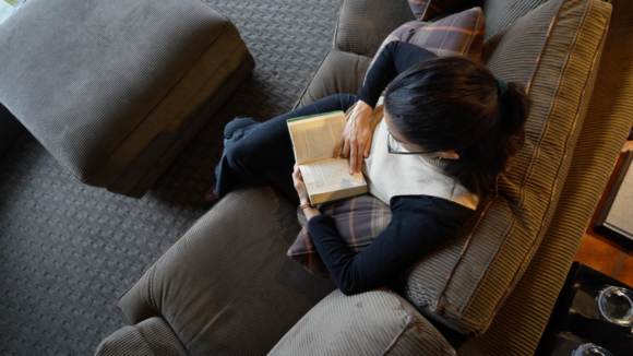 THE SINGULAR SUITE_suite4 Woman wearing glasses reads an open book while relaxing on a cushioned armchair, viewed from above.