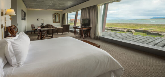 THE SINGULAR SUITE_suite5 Modern hotel suite with a king bed, sitting area, and floor-to-ceiling windows overlooking a grassy shoreline and sea.