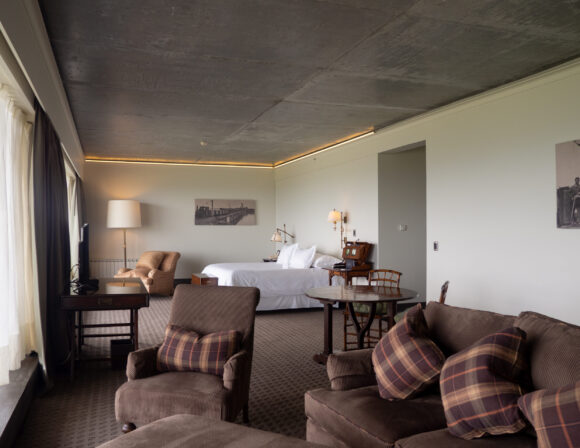 THE SINGULAR SUITE_suite6 Spacious hotel suite with a white bed, plaid sofas, armchair, round table, vintage furnishings, and floor-to-ceiling windows.