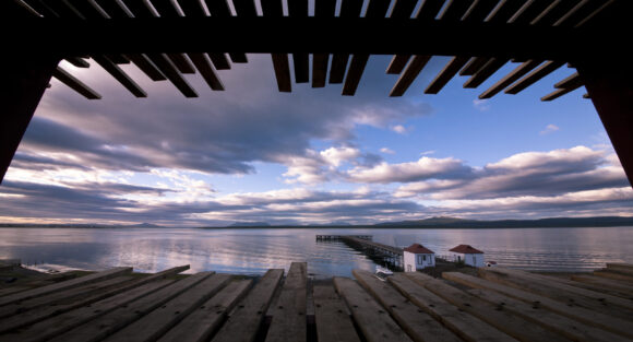 THE SINGULAR SUITE_vista am 312 (1) Sunset over a calm lake and distant mountains, framed by a wooden pergola with a pier and small red-roofed buildings.