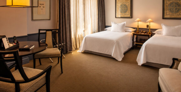 Elegant hotel room with two white beds, a round table and chairs, warm lighting, and framed wall art