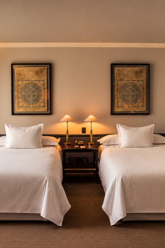 Hotel room with two white twin beds, a shared nightstand, framed artwork, and sunlight through sheer curtains.