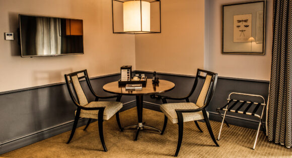 Elegant hotel sitting area with a round table, two upholstered chairs, wall-mounted TV, framed insect art, and pendant light.