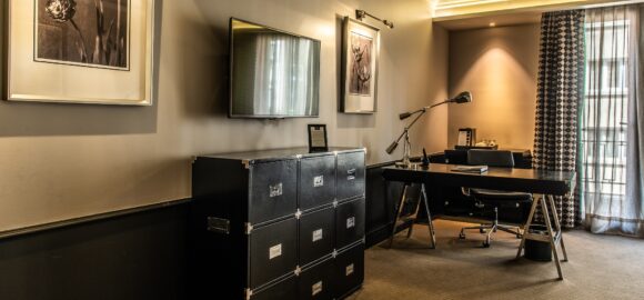 THE SINGULAR TWIN WITH BALCONY_DSC_6632-HDR Elegant hotel room workspace with black desk, filing cabinet, wall-mounted TV, framed floral art, and curtained window.