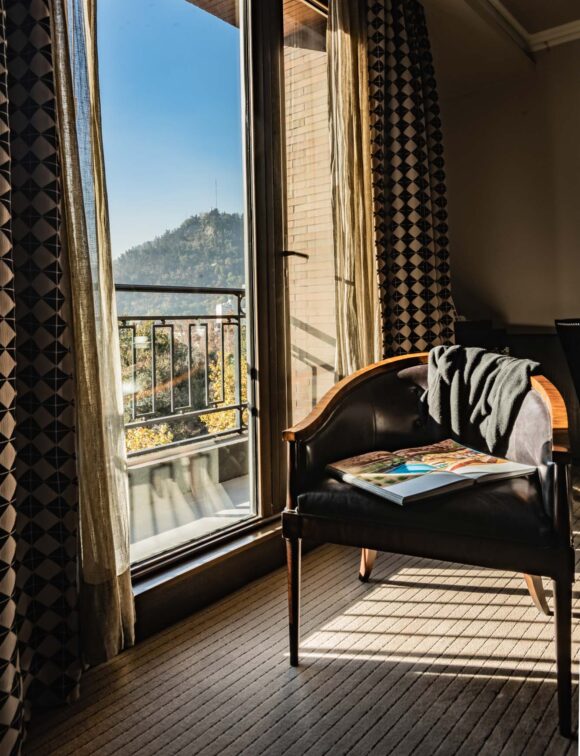 THE SINGULAR TWIN WITH BALCONY_The Singular Suite (25) Sunlit reading nook with an open book on a leather chair beside a balcony window overlooking a wooded mountain.