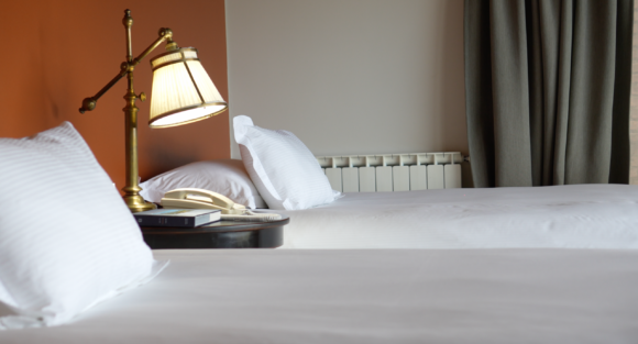 Neatly made hotel bed with white pillows, brass reading lamp, bedside phone and book, orange accent wall, and gray curtains.