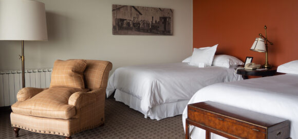 Hotel room with two white beds, a tan armchair, bedside lamp, wooden luggage bench, and rust-red accent wall.