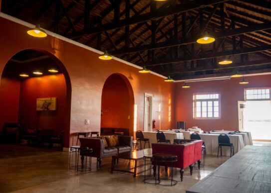 OLYMPUS DIGITAL CAMERA Warmly lit event hall with exposed timber rafters, arched walls, lounge seating, and rows of banquet tables by large windows.