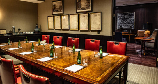 Elegante sala de conferencias con una mesa de madera pulida, sillas de cuero rojo, arte enmarcado y suministros para reuniones.