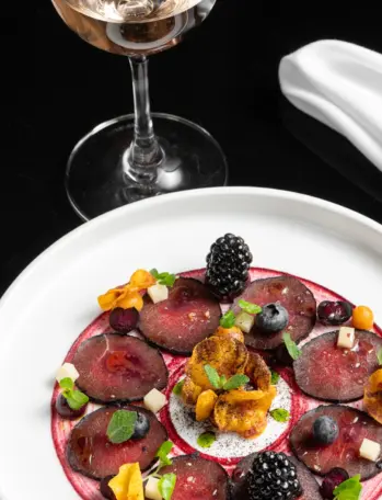 Fine-dining plate of sliced beetroot with berries, crisp garnishes, herbs, and berry sauce beside a wine glass.