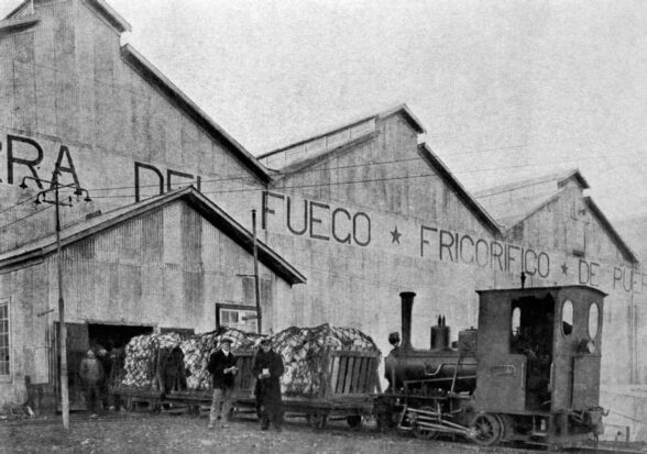 Black-and-white photo of a small locomotive and workers beside a railcar loaded with bundled cargo outside large warehouses.