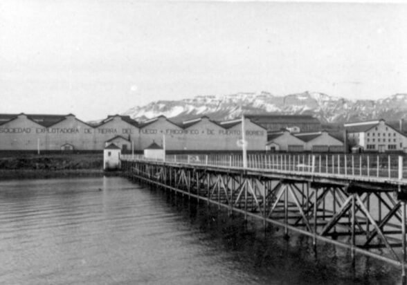 Historic black-and-white view of a wooden pier leading to industrial buildings in Tierra del Fuego, with snowy mountains beyond.