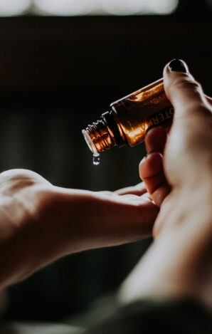Person tilts an amber essential oil bottle, letting a drop fall into their open palm against a dark background.