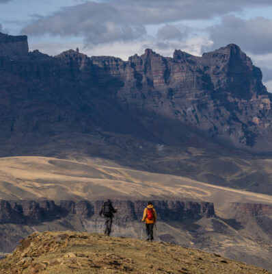 Two hikers stand on a rocky ridge overlooking a vast mountain range beneath cloudy skies.