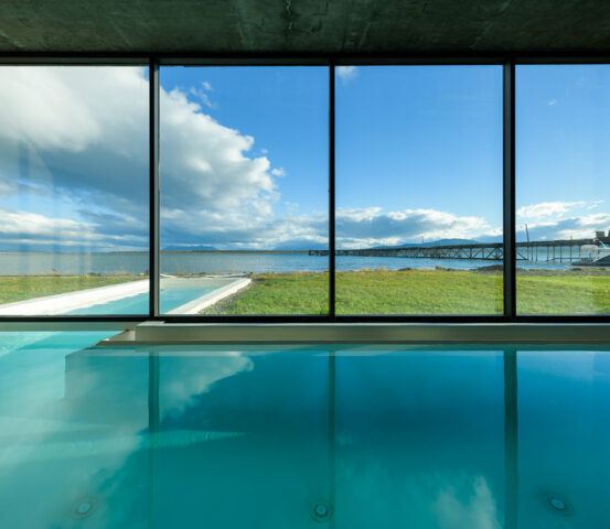 expwellspat_web_HQ Indoor swimming pool overlooking a coastal bay, green lawn, pier, and boat through expansive floor-to-ceiling windows.