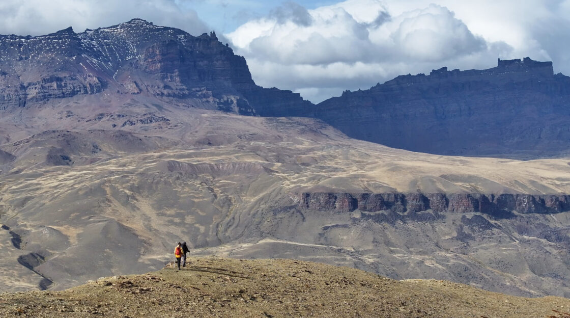 Captura de pantalla Two hikers traverse a barren alpine ridge beneath towering, snow-dusted mountains and low clouds.
