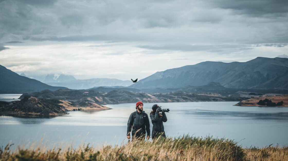 hon4 Two hikers photograph a bird flying over a mountain lake beneath a cloudy sky.