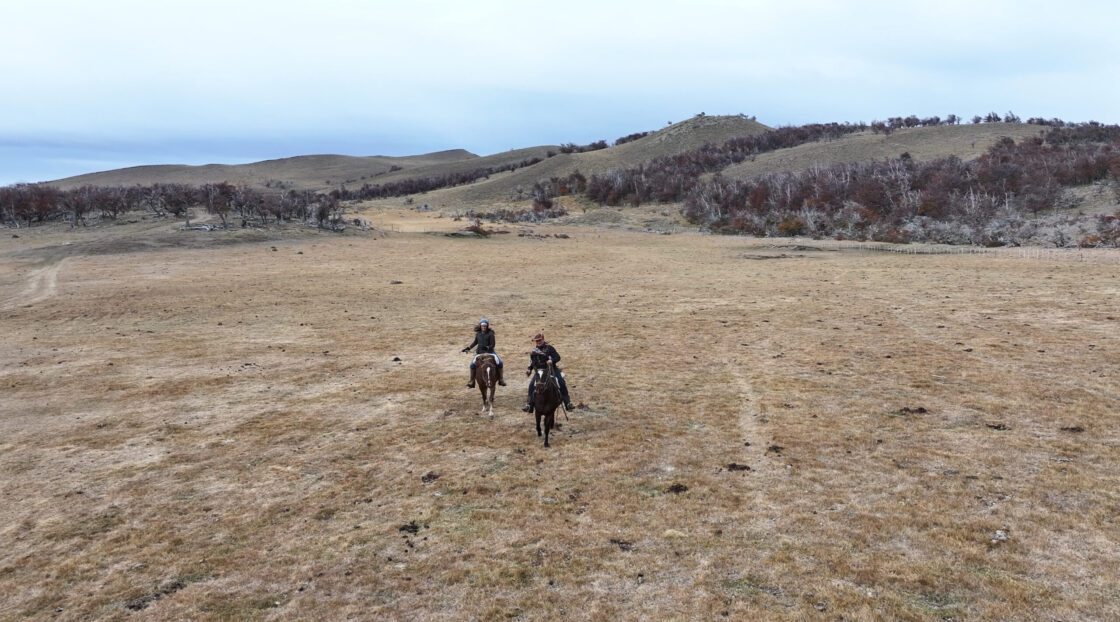 Captura de pantalla Two riders on horseback cross a dry grassland beneath rolling hills and sparse trees.