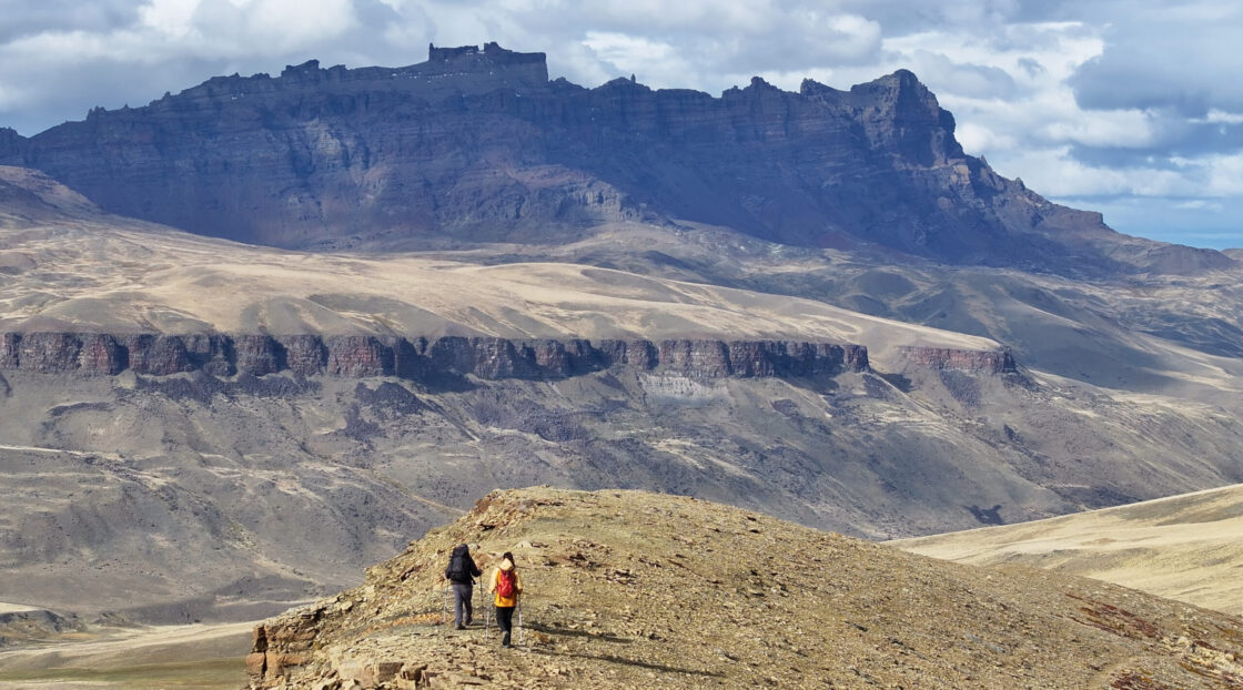 Captura de pantalla Two hikers with trekking poles cross a rocky ridge beneath a vast, layered mountain range and cloudy sky.