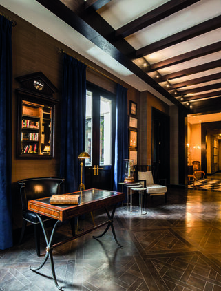 Elegant dark wood-paneled library with a writing desk, bookshelves, tall windows, and a warmly lit adjoining room.