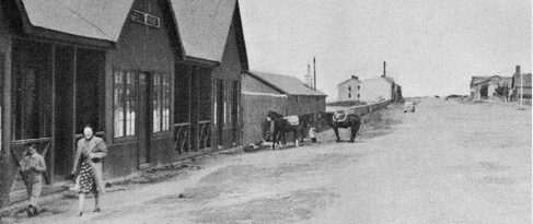 Black-and-white photo of wooden storefronts, pedestrians, and horses on a quiet rural street.