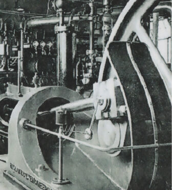 Black-and-white archival photo of a large industrial steam engine with flywheel, piston, and connecting rods in a factory