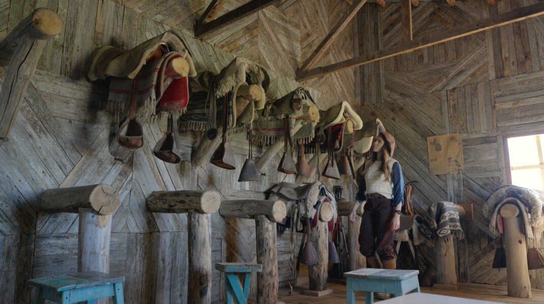 Screenshot Woman examines traditional saddles and riding gear hanging inside a rustic wooden barn.