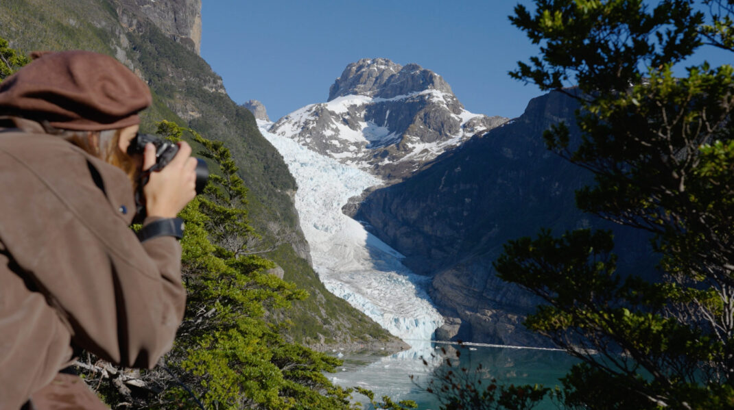 Screenshot Hiker photographs a snow-covered mountain glacier descending into a turquoise lake in a rugged valley.